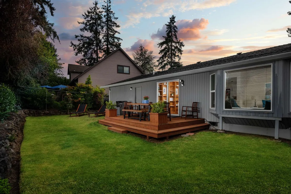 This exterior photograph features a spacious wooden deck with a large dining table set up for outdoor meals, surrounded by lush green lawn and mature trees. The deck is well-maintained and invites relaxation or group gatherings, with ample space for both kids and pets to move around safely. The view showcases the tranquil, private aspect of the backyard, reinforcing its suitability for alfresco living.
