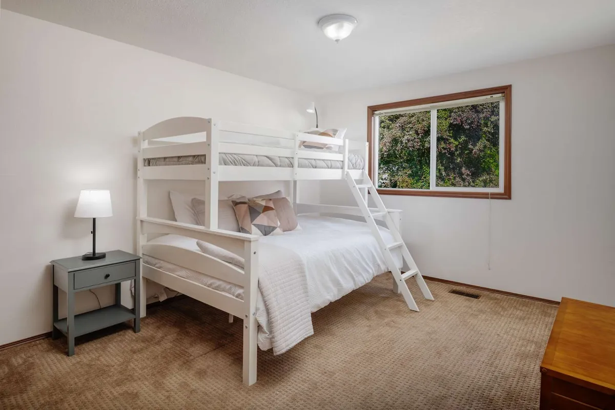 A cozy bunk room is depicted, complete with a bunk bed, window, and nightstand with a lamp. The inviting space feels perfect for sleepovers, reading, and relaxing, with subtle suggestions of movie or game nights.