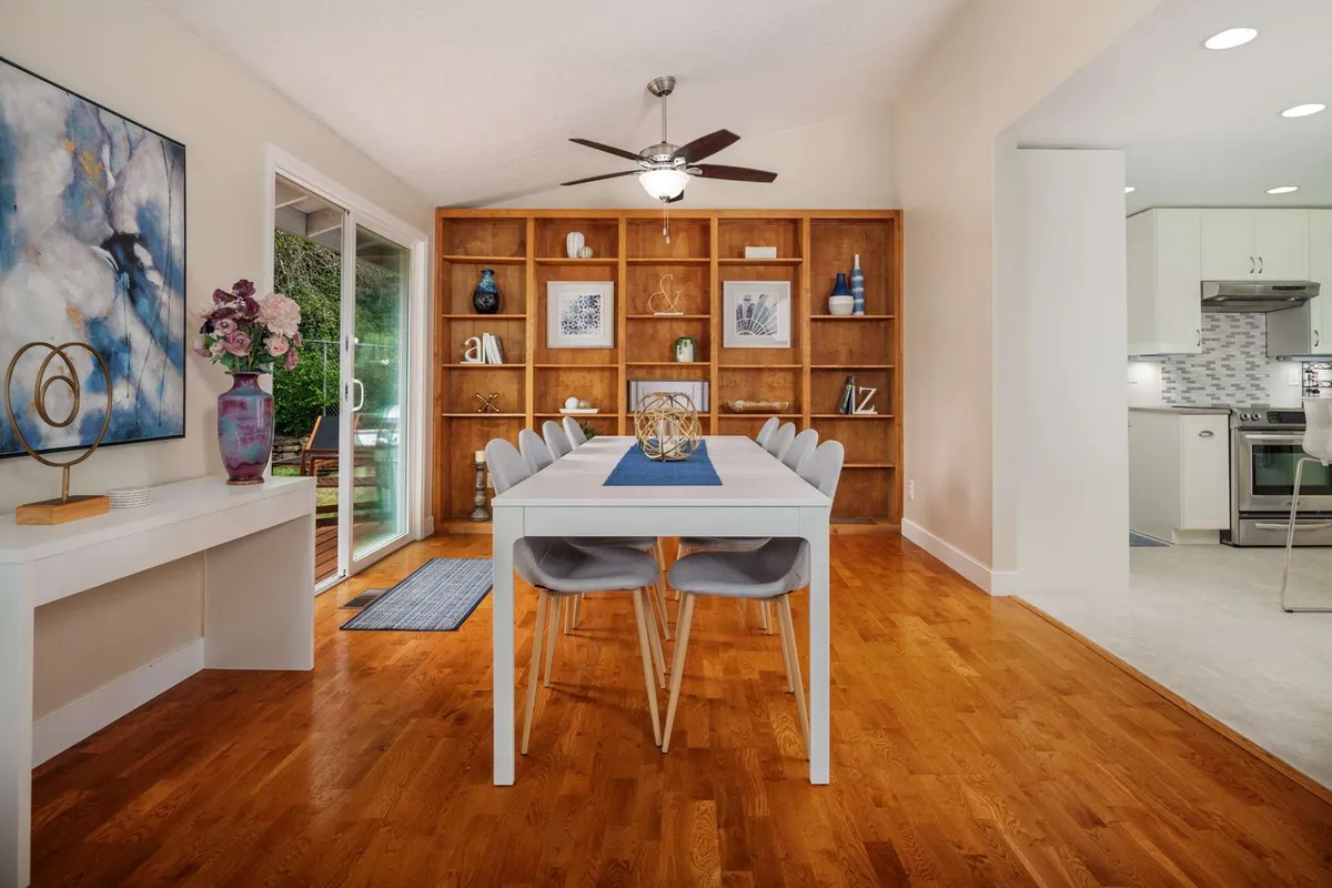 A welcoming, contemporary dining space featuring a large table, wooden shelving, and artwork. The area is set up for gatherings, with a vase of flowers as a centerpiece, suggesting communal meals, celebrations, and group interactions.