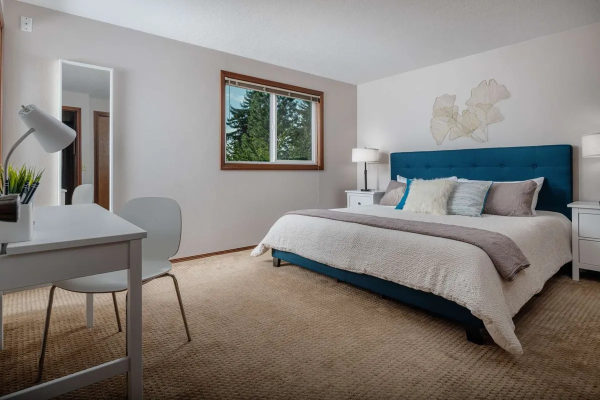 A serene bedroom with a blue upholstered bed, crisp white bedding, a window that frames lush greenery, and a small desk with a lamp. The scene suggests a tranquil atmosphere, ideal for restful nights and quiet moments of work or reflection.