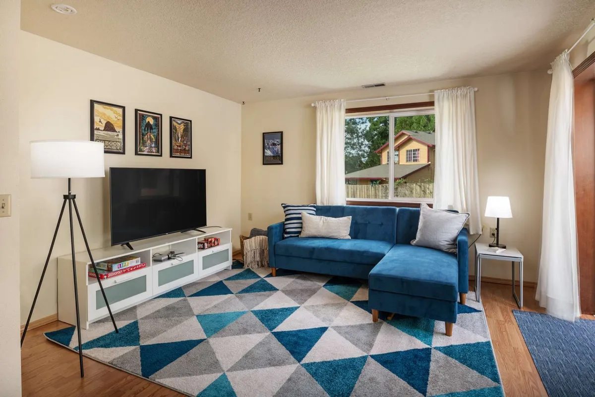 A cozy living area with a blue sectional sofa facing a television, geometric area rug, and contemporary wall art. The ambiance is warm and inviting, ideal for relaxing, watching TV, or playing board games together.