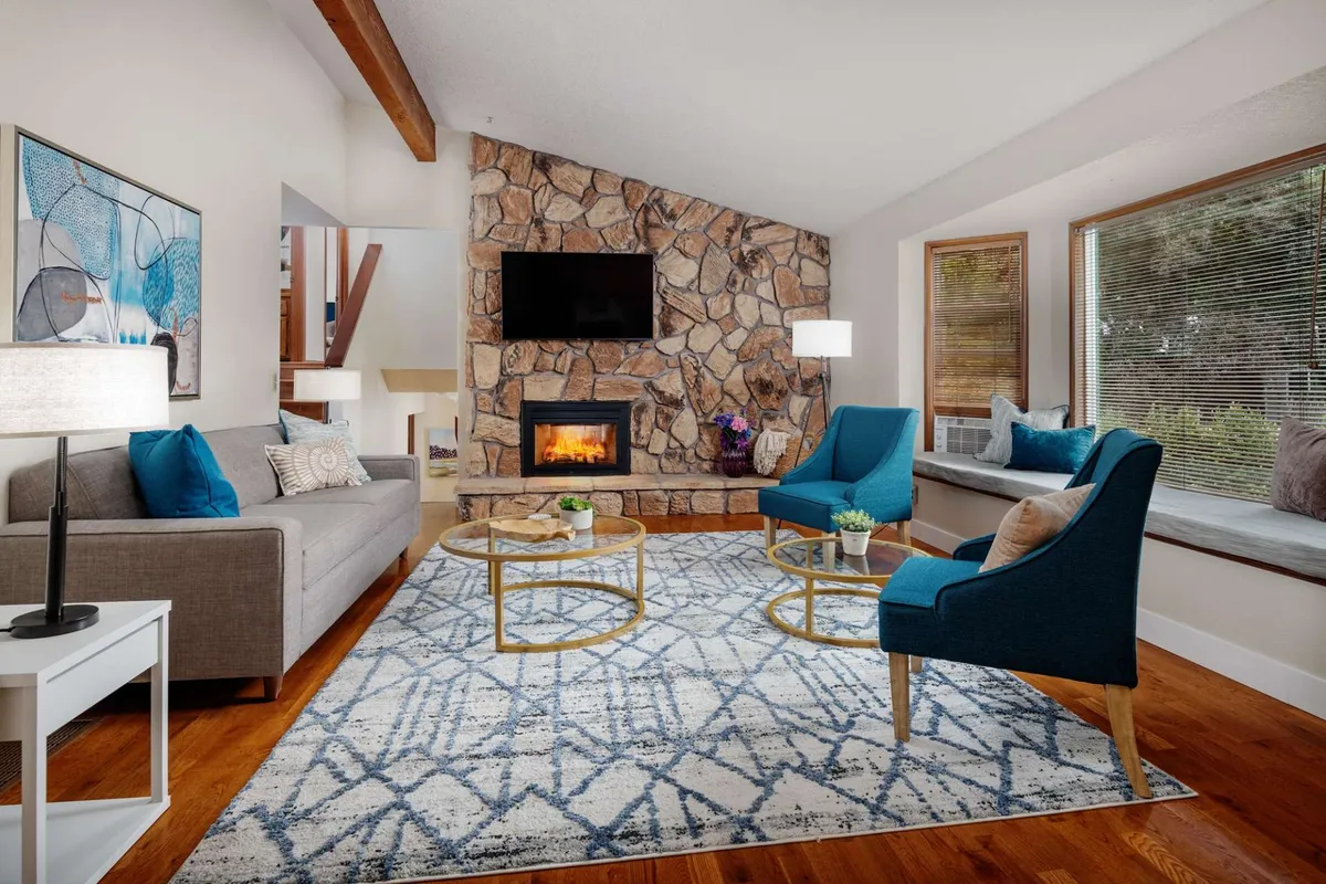 A living room scene featuring a stone fireplace, blue accent chairs, and artwork on the wall. The bright, welcoming space is arranged for relaxing, socializing, and, with ample seating, enjoying TV or board games together.