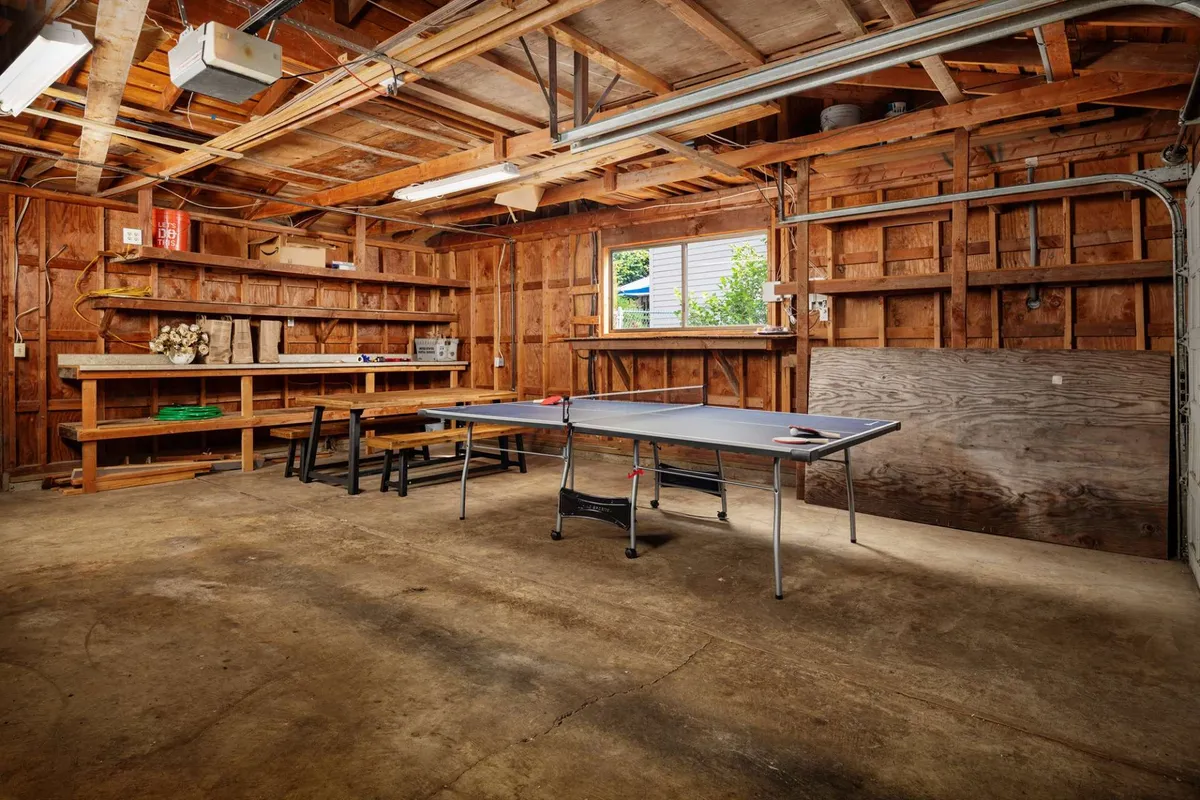 This image showcases a garage space featuring a ping pong table positioned in the center. Shelves, wooden beams, and a window provide a relaxed, informal setting perfect for friendly matches or casual gatherings. The open, versatile nature of the space hints at its use for various fun activities.