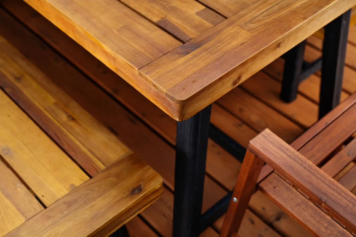 The image centers on a rustic wooden table and bench, highlighting the fine grain and craftsmanship of the outdoor furniture. The intimate setting suggests casual family meals, celebrations, or peaceful brunches under open skies. Though the wider yard isn't visible, the furniture itself evokes the section's communal, celebratory outdoor spirit.